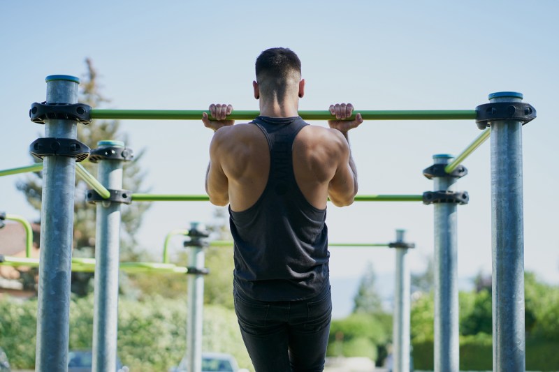 A man in black sleeveless top doing pull ups with a bar at a park.