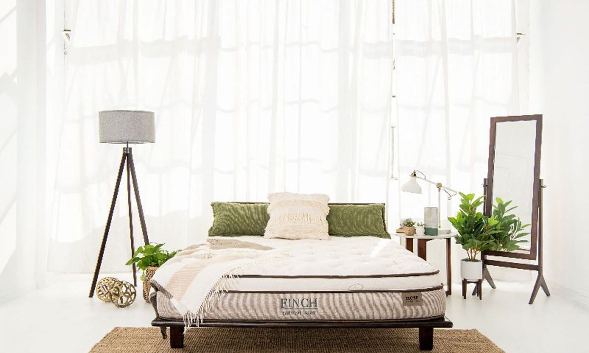 A Finch mattress on a bedframe beside a lamp, bedside table, potted plants, and mirror.