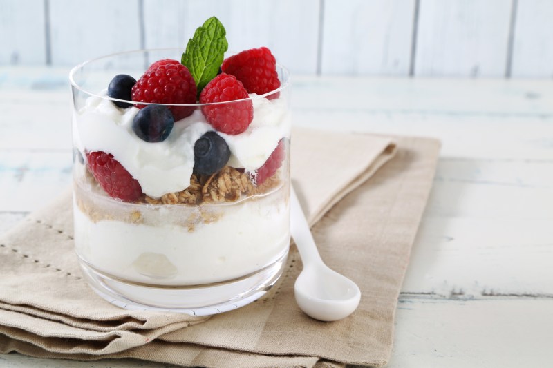 Un vaso de parfait de yogur con bayas y granola junto a una cuchara de plástico sobre un paño marrón. 