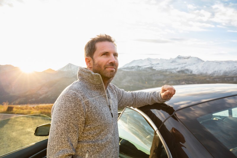 A man in gray half-zip pullover sweater placing his arm on his car in the outdoors.