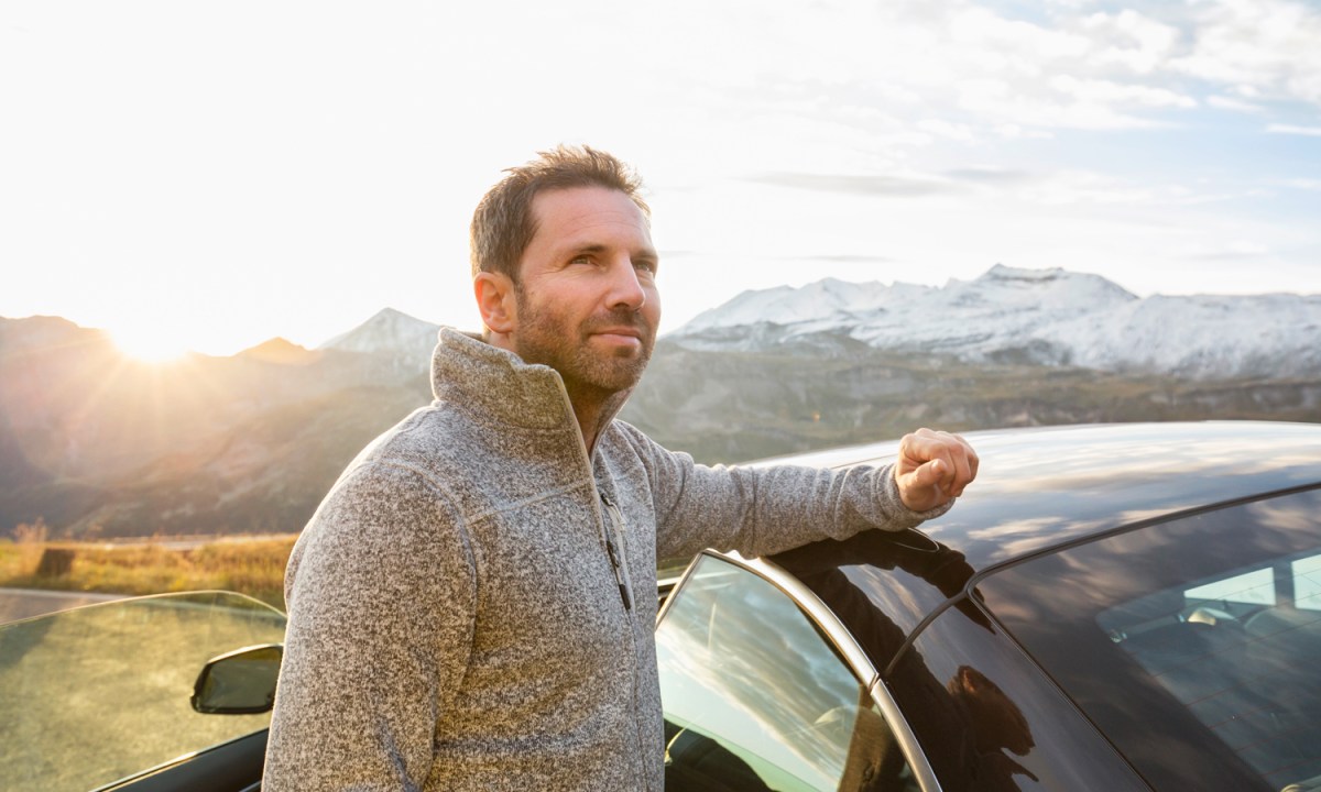 A man in gray half-zip pullover sweater placing his arm on his car in the outdoors.