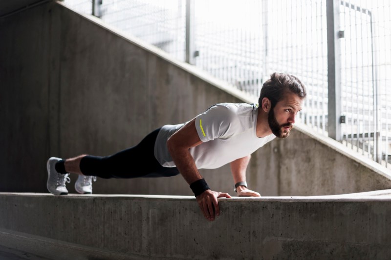 A man in white shirt, black jogging pants, and white shoes doing push-ups on a concrete platform.