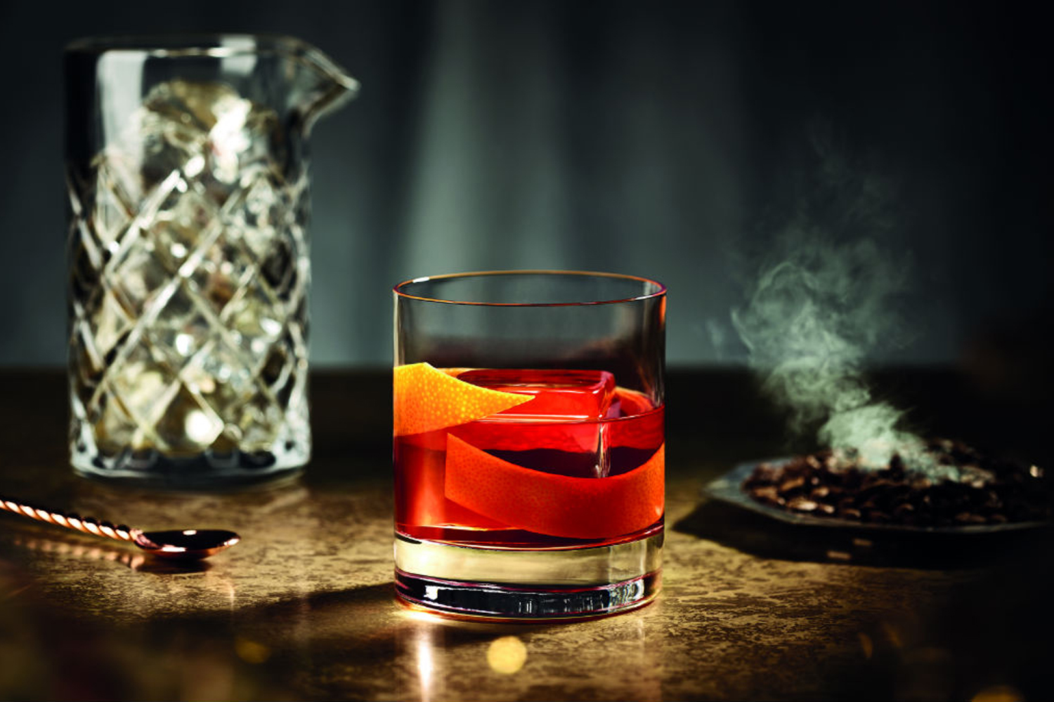 A Jitney Negroni garnished with orange on a table with spoon, pitcher with ice, and cigarette tray in the background.