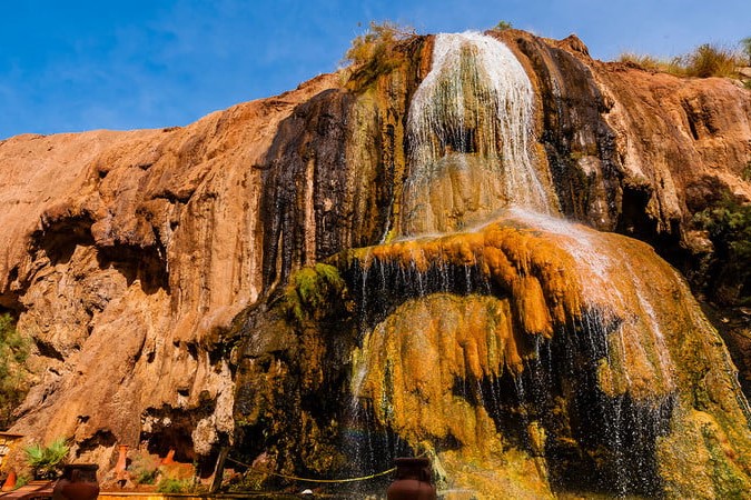 A hot springs area in Jordan, surrounded by a brown cliff. There is a waterfall trickling down from the top of the cliff to the base of the hot springs area.