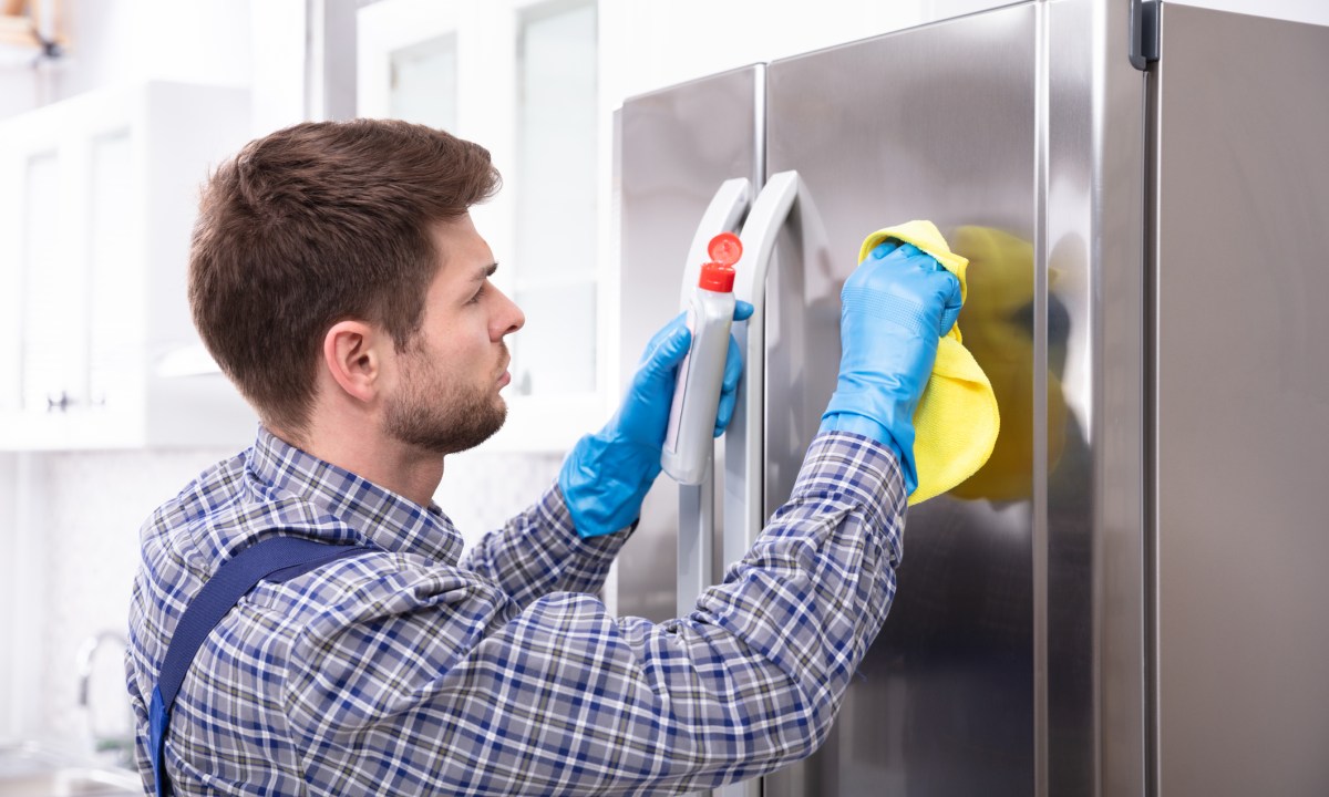 Side View Of Man Cleaning Refrigerator With Cloth And Bottle.