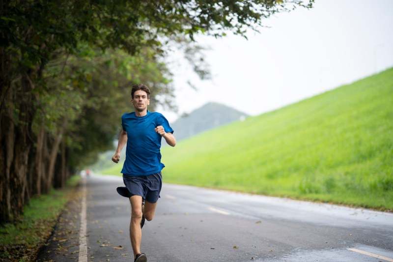 A man in a blue shirt and navy blue shorts jogging on a road with trees and a lawn in the background. 