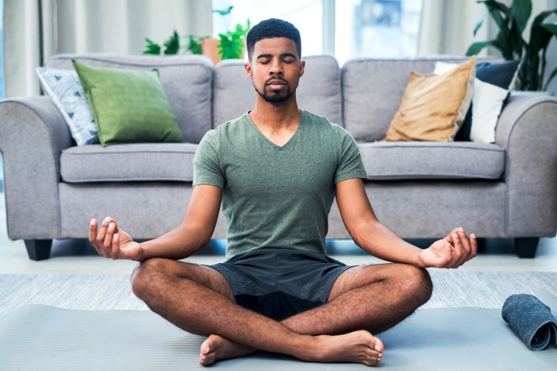 African american man in green shirt and gray shorts meditating on yoga mat in living room.