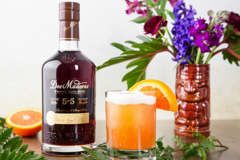 A glass of Pearl Diver cocktail garnished with orange beside a bottle of liquor and flowers along with plant stalks and a sliced orange