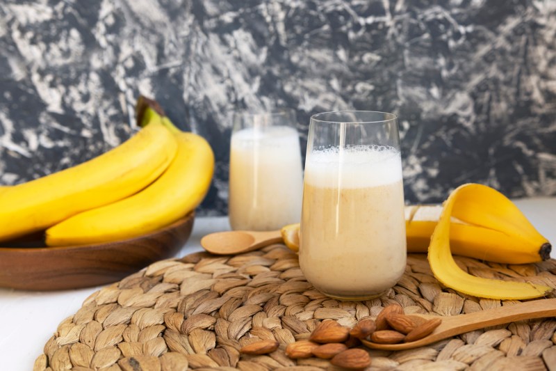 Un batido de plátano con plátanos, nueces, cucharas de madera, posavasos y un vaso de leche.