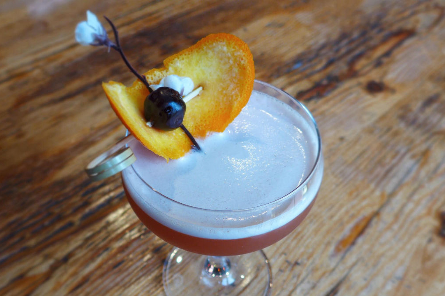 A glass of They Call Him Singapore "The Sling" garnished with orange on wooden table.