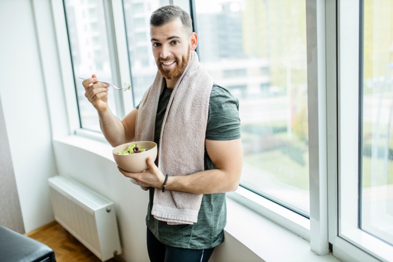 Un hombre con una toalla sobre su hombro comiendo una ensalada después de hacer ejercicio en casa.