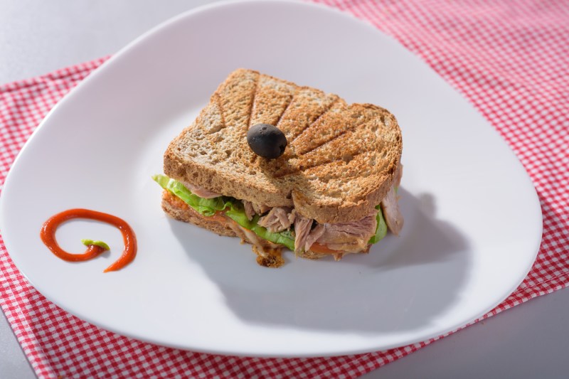 Un sándwich tostado con atún y verduras y salsa en un plato sobre tela roja y blanca.