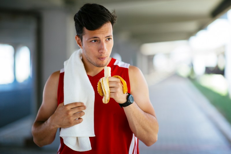 A man with a towel over his shoulder eating a banana.