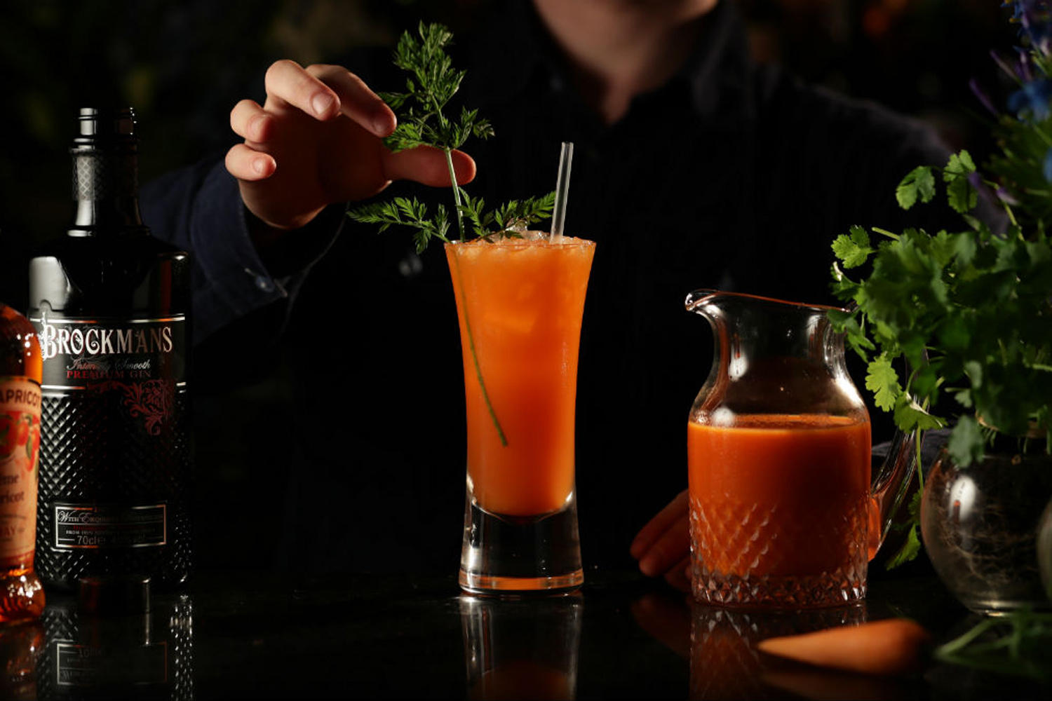 A bartender garnishing a What's Up Brock Cocktail coriander leaves beside a pitcher of What's Up Brock cocktail and a bottle of Brockman's gin.