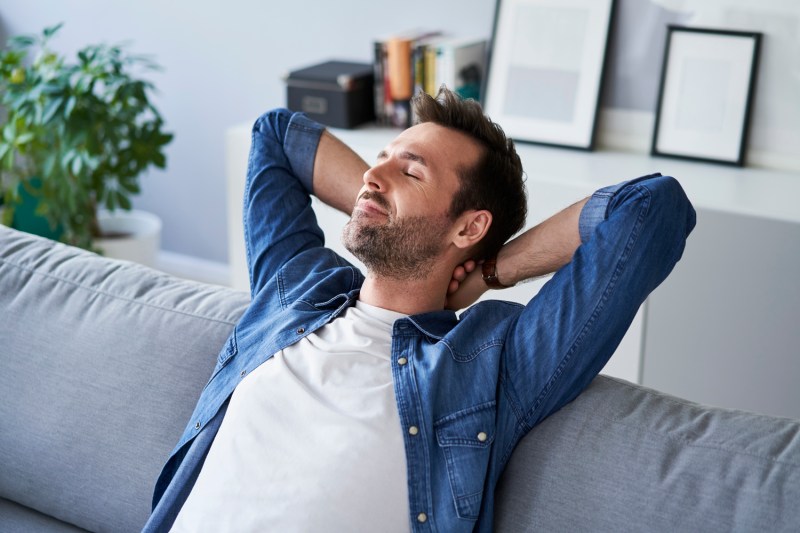 A man, his eyes closed, resting on a gray sofa, his hands behind his neck.