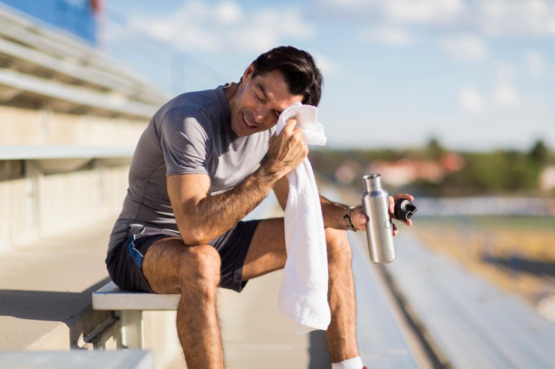 A man sitting on the bleachers holding a water bottle while wiping his brow with a white towel.