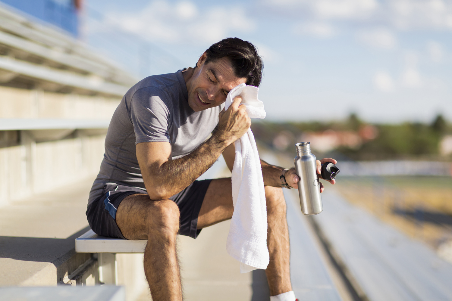 A man sitting on the bleachers holding a water bottle while wiping his brow with a white towel.