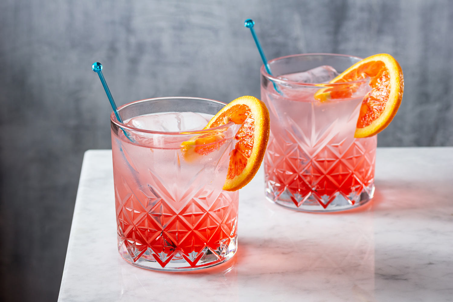 Two ice-cold glasses of gin with blood orange garnish with cocktail sticks on a table.
