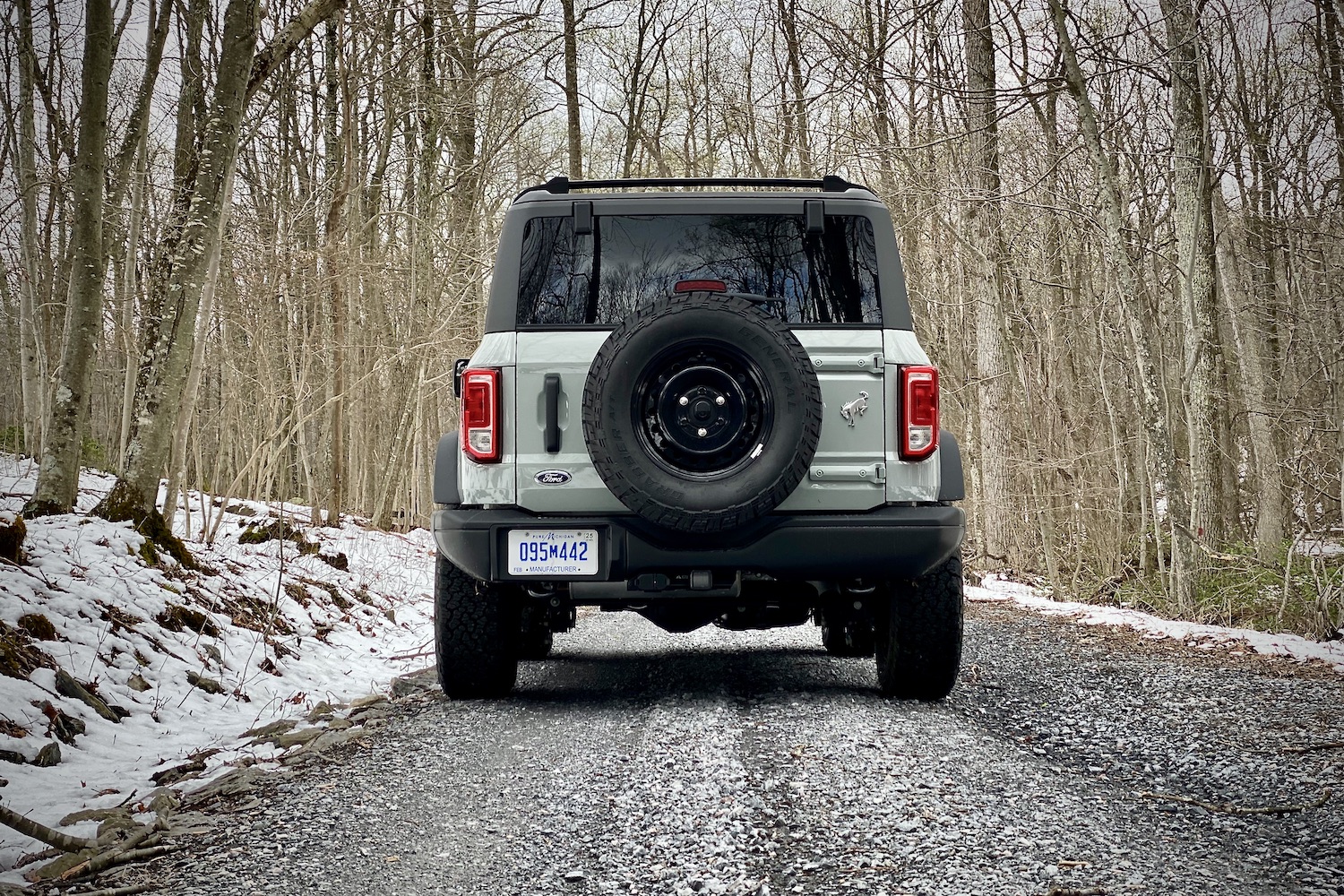 Rear end of 2021 Ford Bronco on a gravel trail with trees and snow in the back.