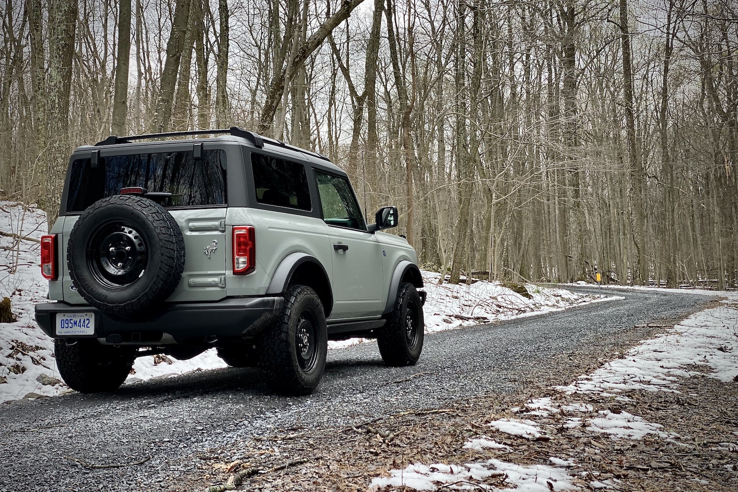 Rear end angle of 2021 Ford Bronco from passenger side on a gravel trail with trees and snow in the back.