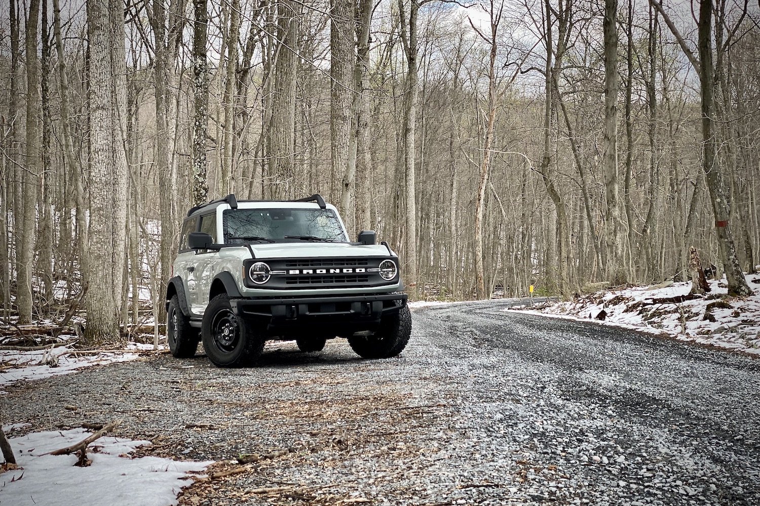 Front end angle of 2021 Ford Bronco from passenger side on a gravel trail with trees in the back on a cloudy day.