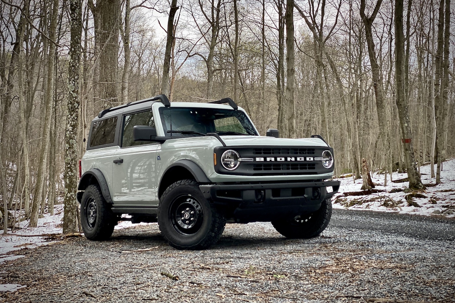 Front end angle of 2021 Ford Bronco on a gravel trail with trees in the back on a snowy cloudy day.