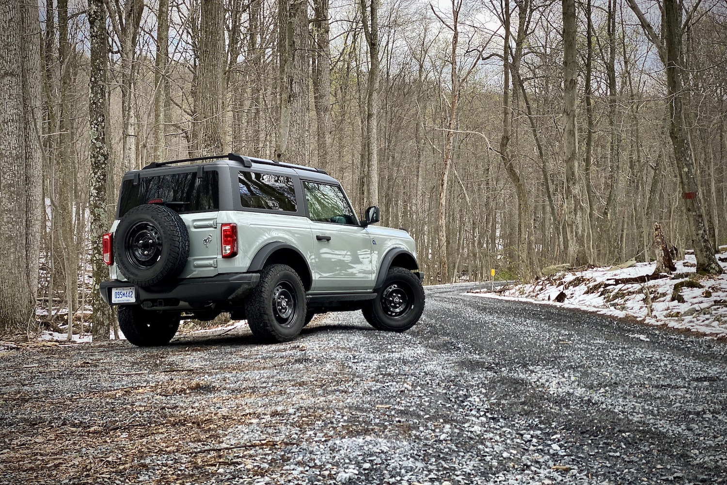 Rear end angle of 2021 Ford Bronco on a gravel trail with trees and snow in the back on a gloomy day.