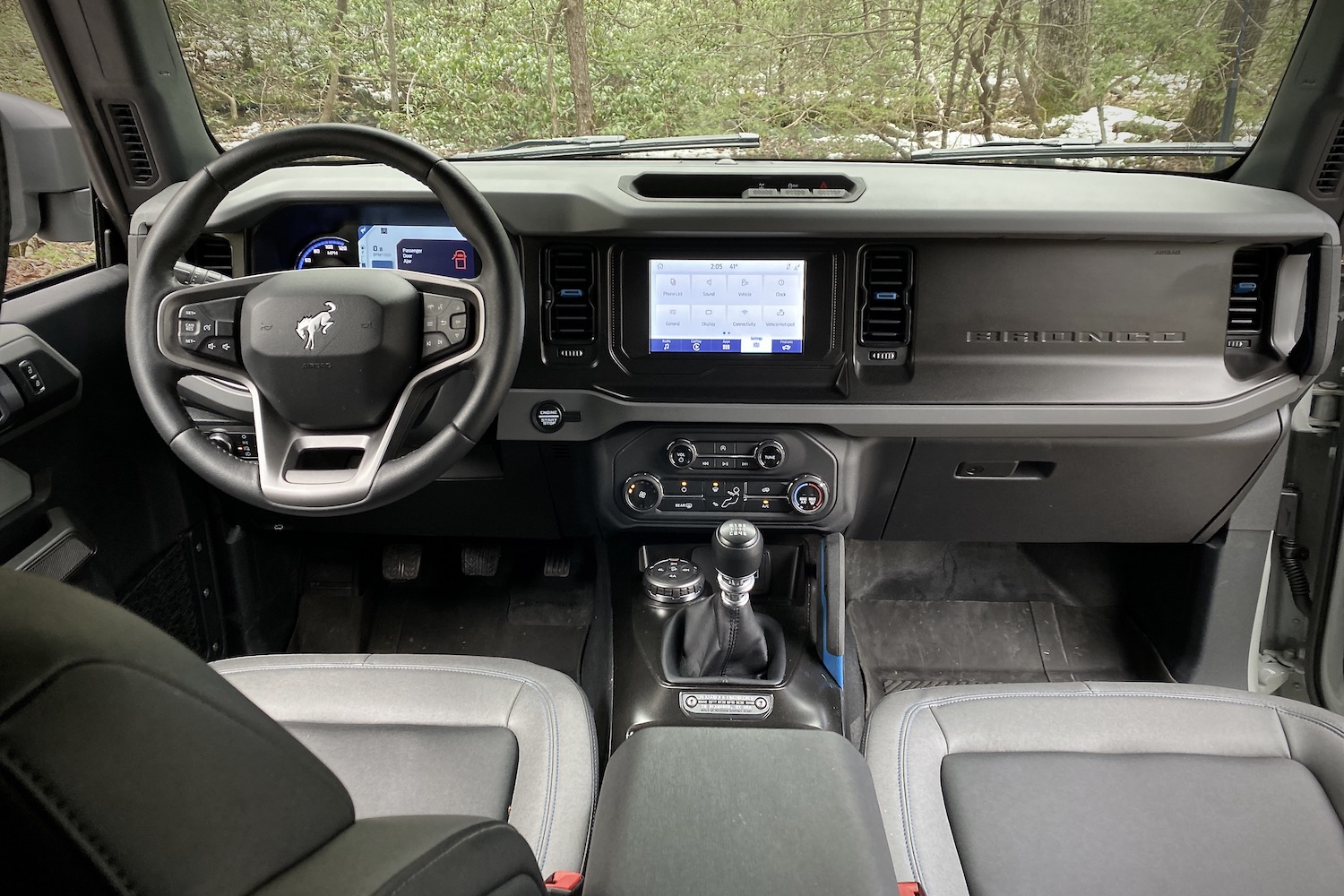 Steering wheel, dashboard, and center console in the 2021 Ford Bronco from the back seats with trees in the back.