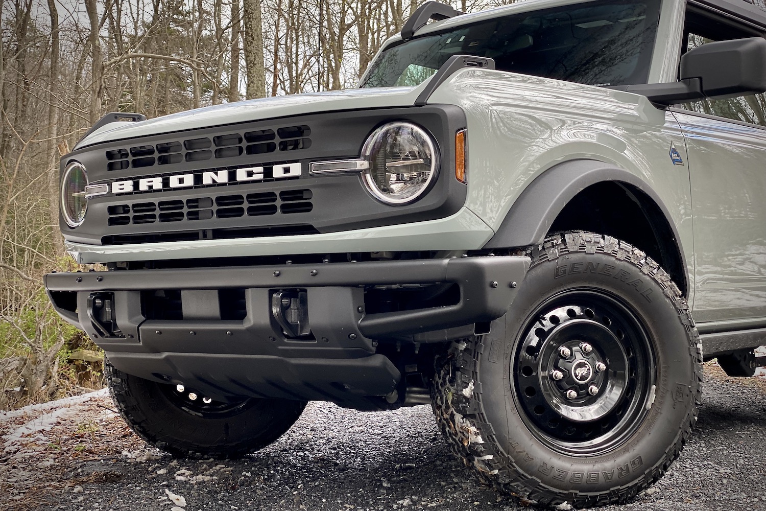 Close up of 2021 Ford Bronco front end from driver's side angle on a gravel trail with trees in the back.