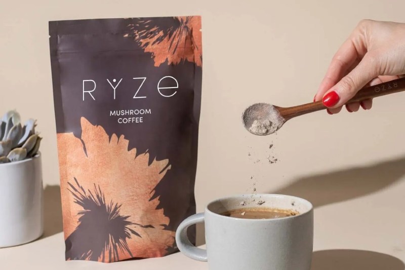 Ranka įmeta Ryze grybų kavos gabalėlį į taurę su Ryze paketu už jos