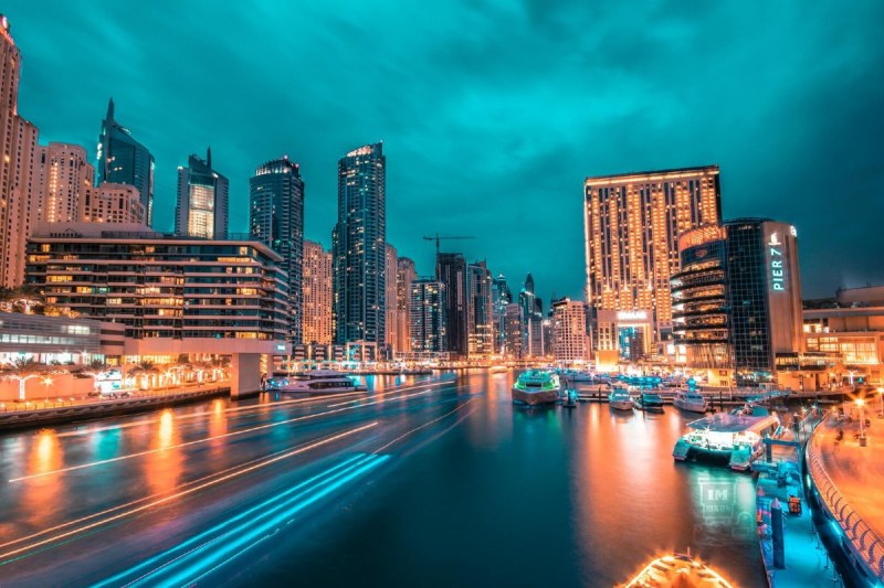 A city nightscape featuring a river in the foreground, and lit streets and buildings underneath an aqua blue sky