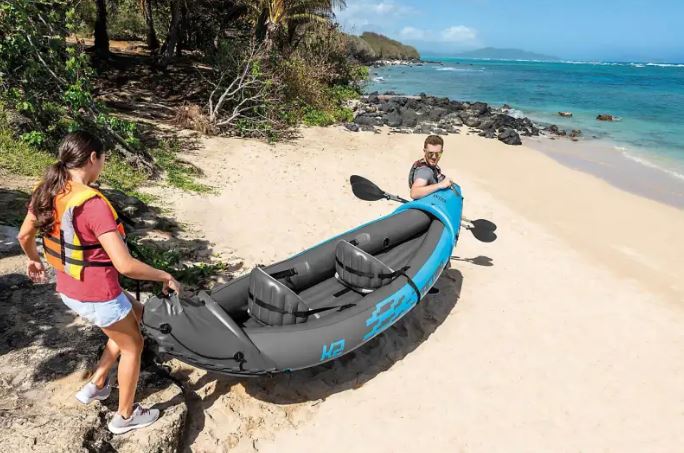 Two people carry a blue inflatable kayak onto a tropical beach.