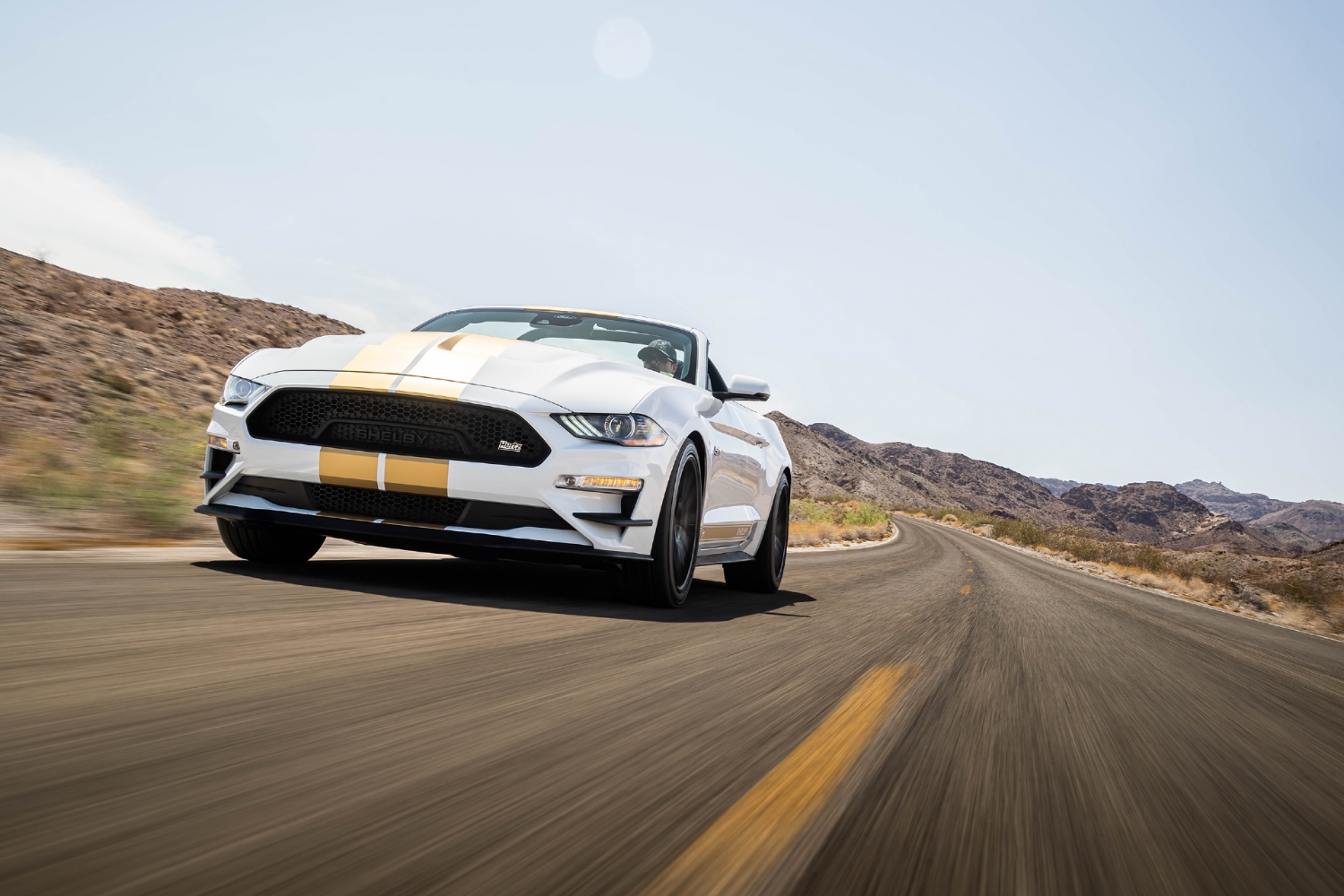 White and gold 2022 Shelby GT-H Convertible driving on a two-lane road in the desert with blue skies.