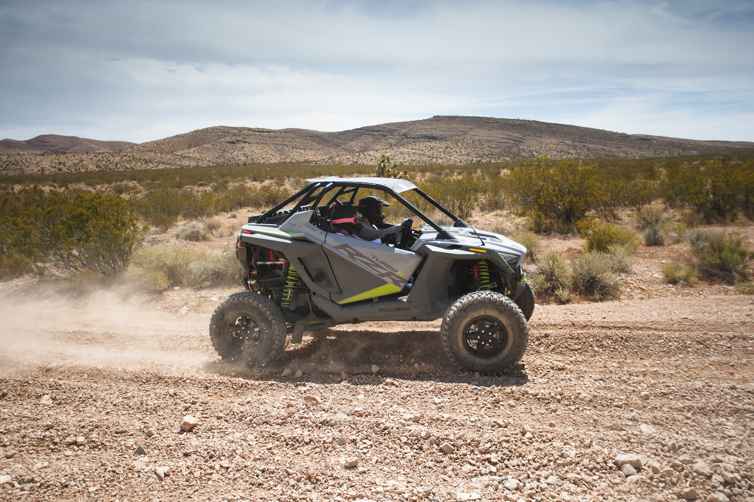 Closeup of side angle of Polaris RZR Turbo R on a dirt trail in the desert with mountains in the background.