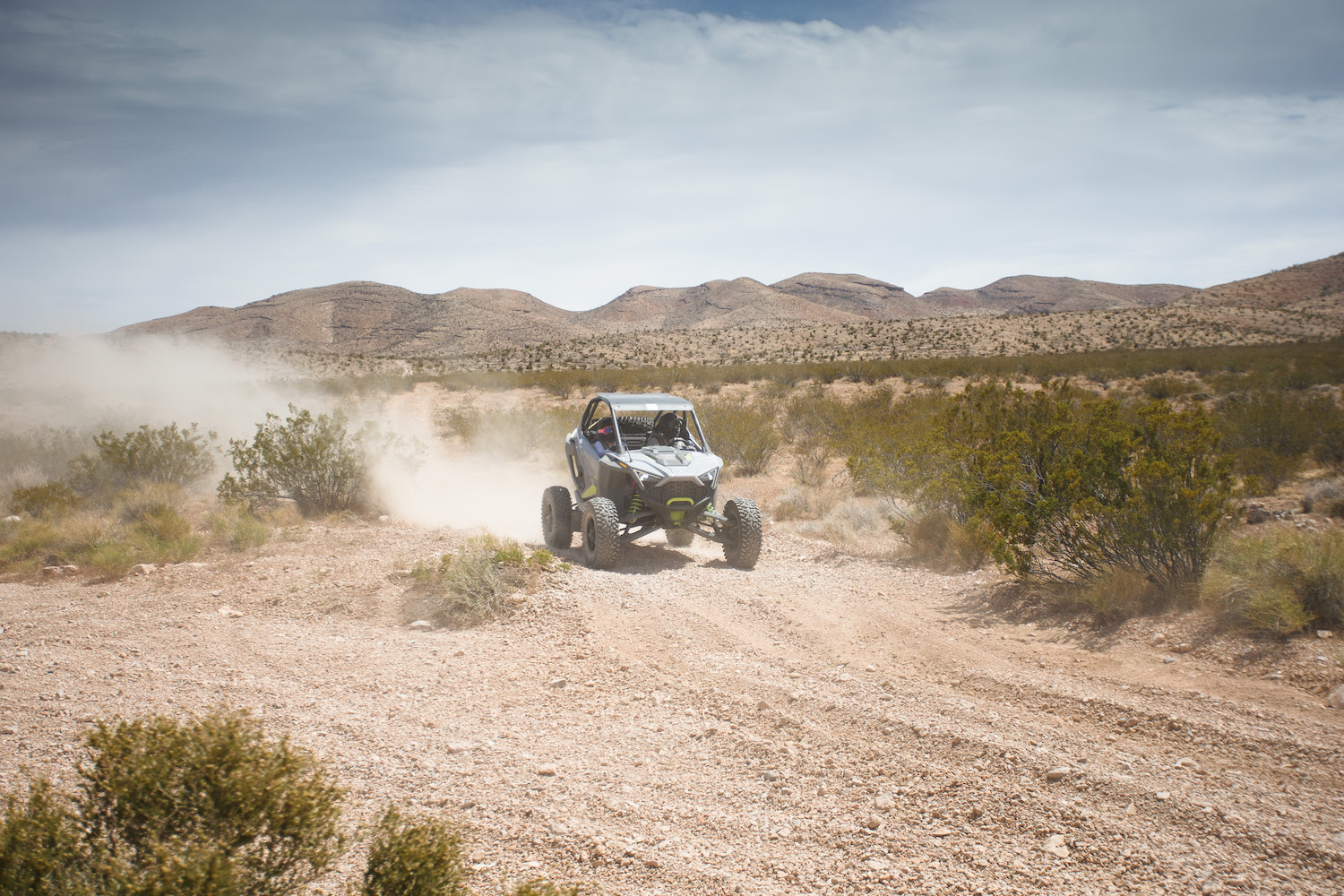 Polaris RZR Turbo R sliding on a dirt path in the desert with mountains and dust in the background.