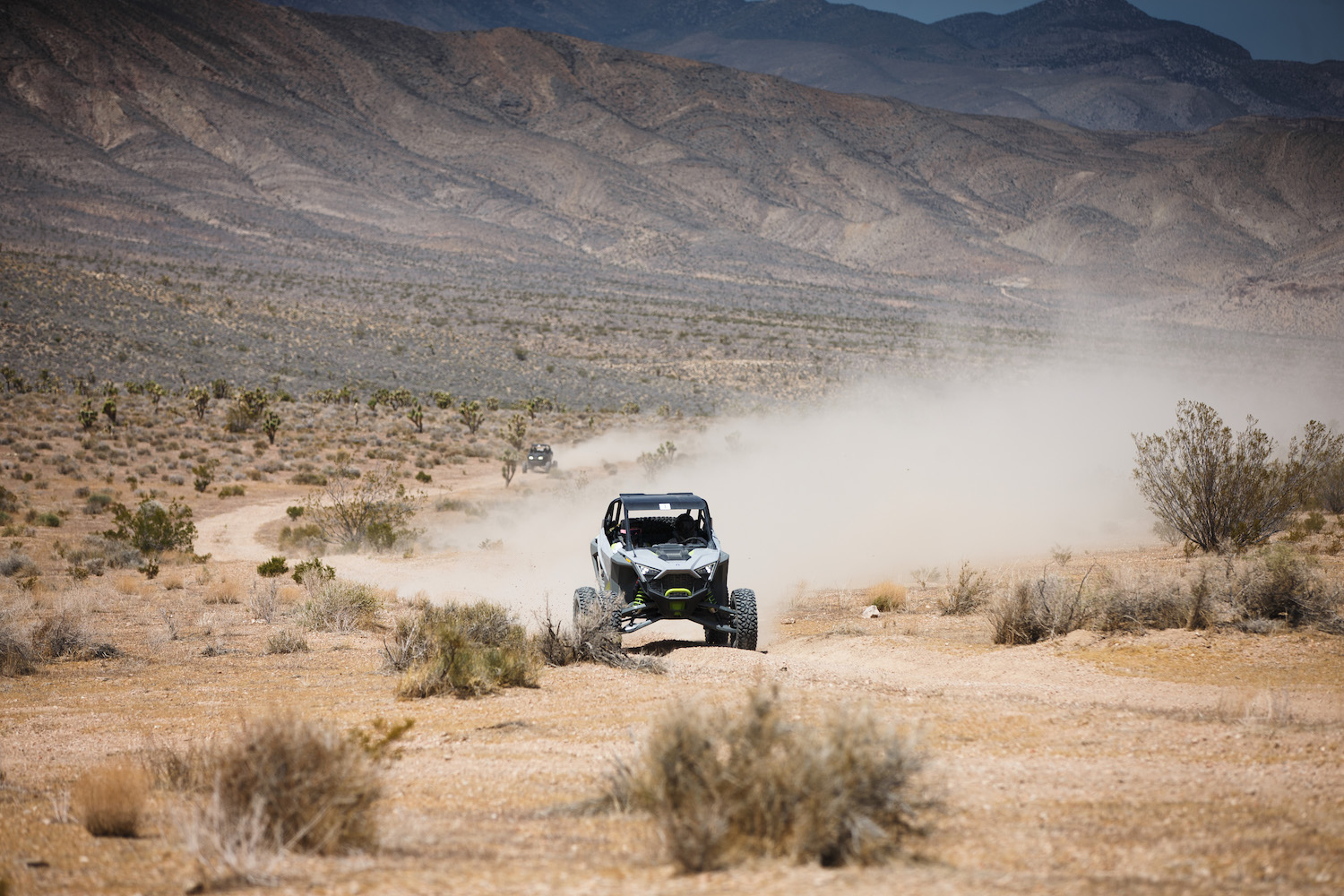 Far away shot of front end of Polaris RZR Turbo R driving through the desert with a dust cloud on a dirt path.