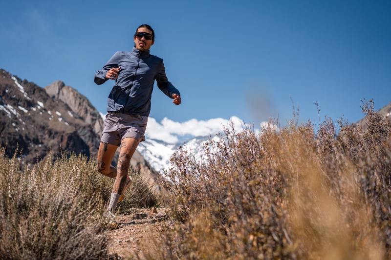 A man in sunglasses runs downhill past a close up image of heather.