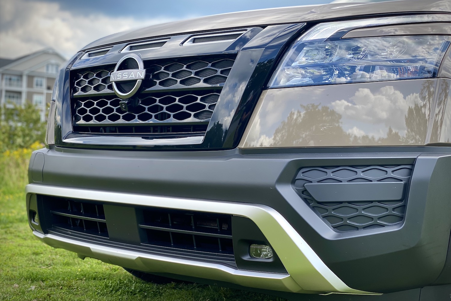 Close up of 2023 Nissan Pathfinder Rock Creek front grille and headlight on a grassy field with a hotel in the back.