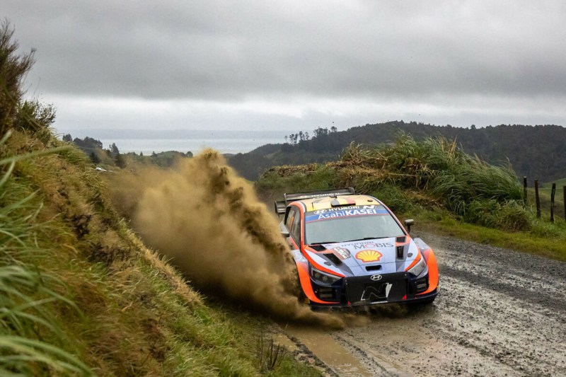 Hyundai i20N WRC rally car driving through thick mud on a dirt road with mountains in the back.
