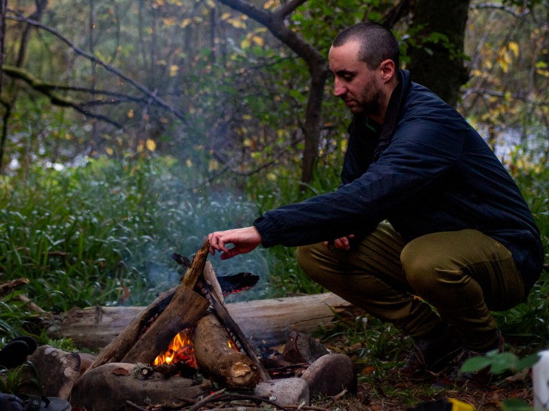 A man puts another log on the campfire wearing Ibex Nomad pants.