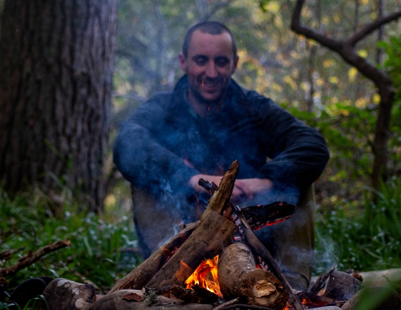 A man sits on the far side of the campfire while smoke rises in front of him.