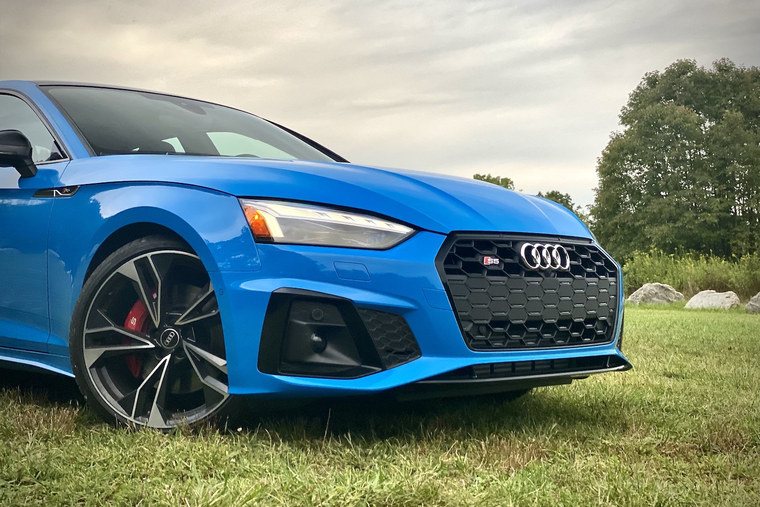 Front end angle of 2023 Audi S5 Sportback from passenger's side in a grassy field during sunset.