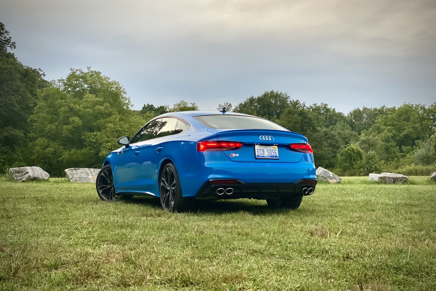Rear end angle of 2023 Audi S5 Sportback from driver's side in a grassy field during a sunset.