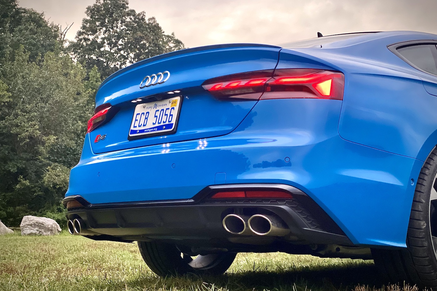 Close up of rear end of 2023 Audi S5 Sportback from below with trees in the back.