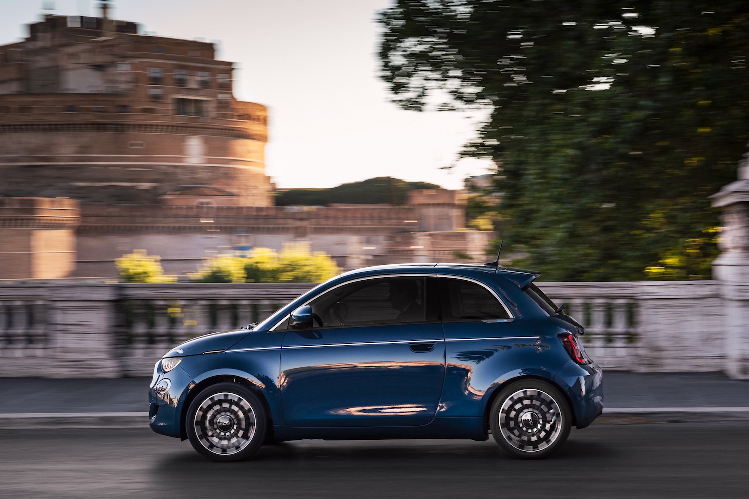 2024 Fiat 500e side profile driving in the city with tall buildings in the back.