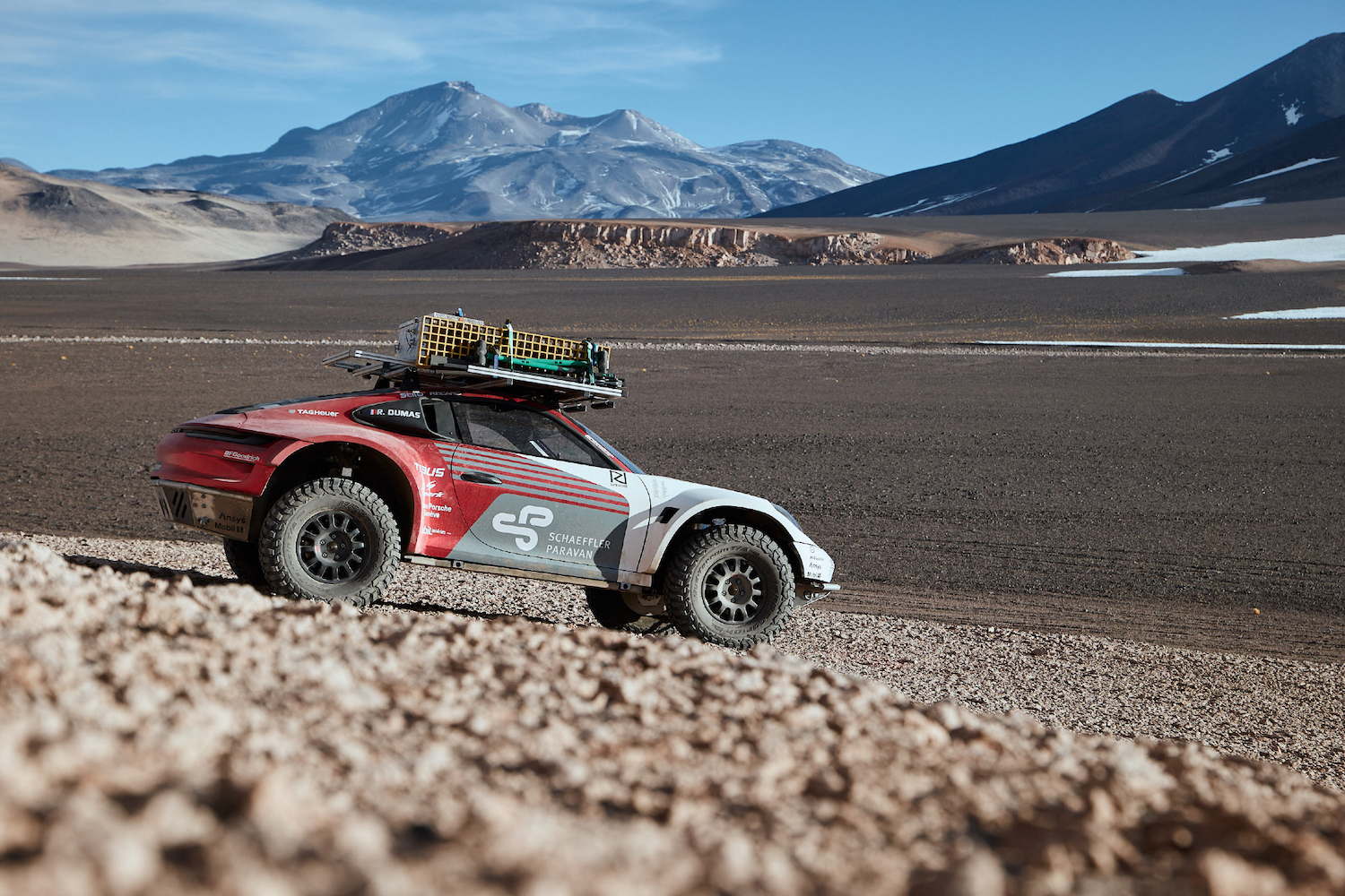 Porsche 911 Dakar Prototype going down a rocky hill with mountains and blue skies in the back.