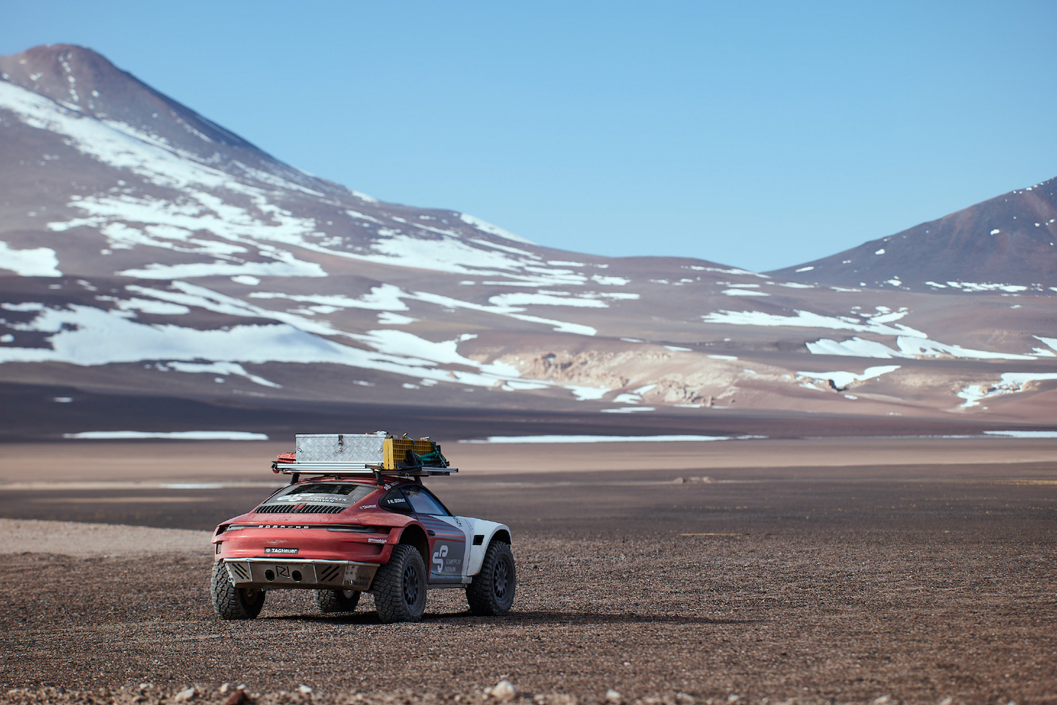 Rear end of Porsche 911 Dakar Prototype driving on rocky terrain with snow on mountains in the back.