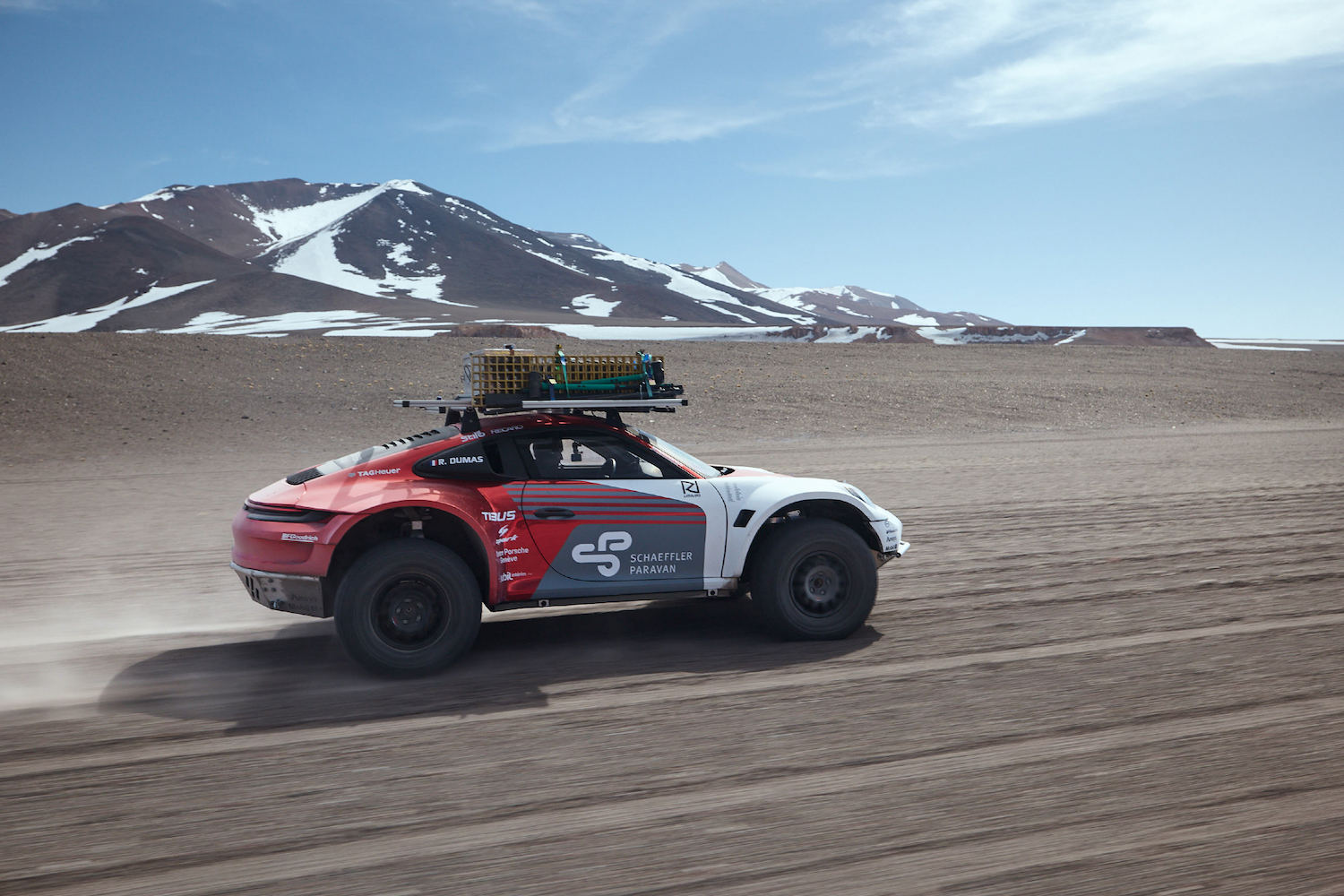 Porsche 911 Dakar Prototype rear end driving fast on rocky terrain with snow-capped mountain in the back.