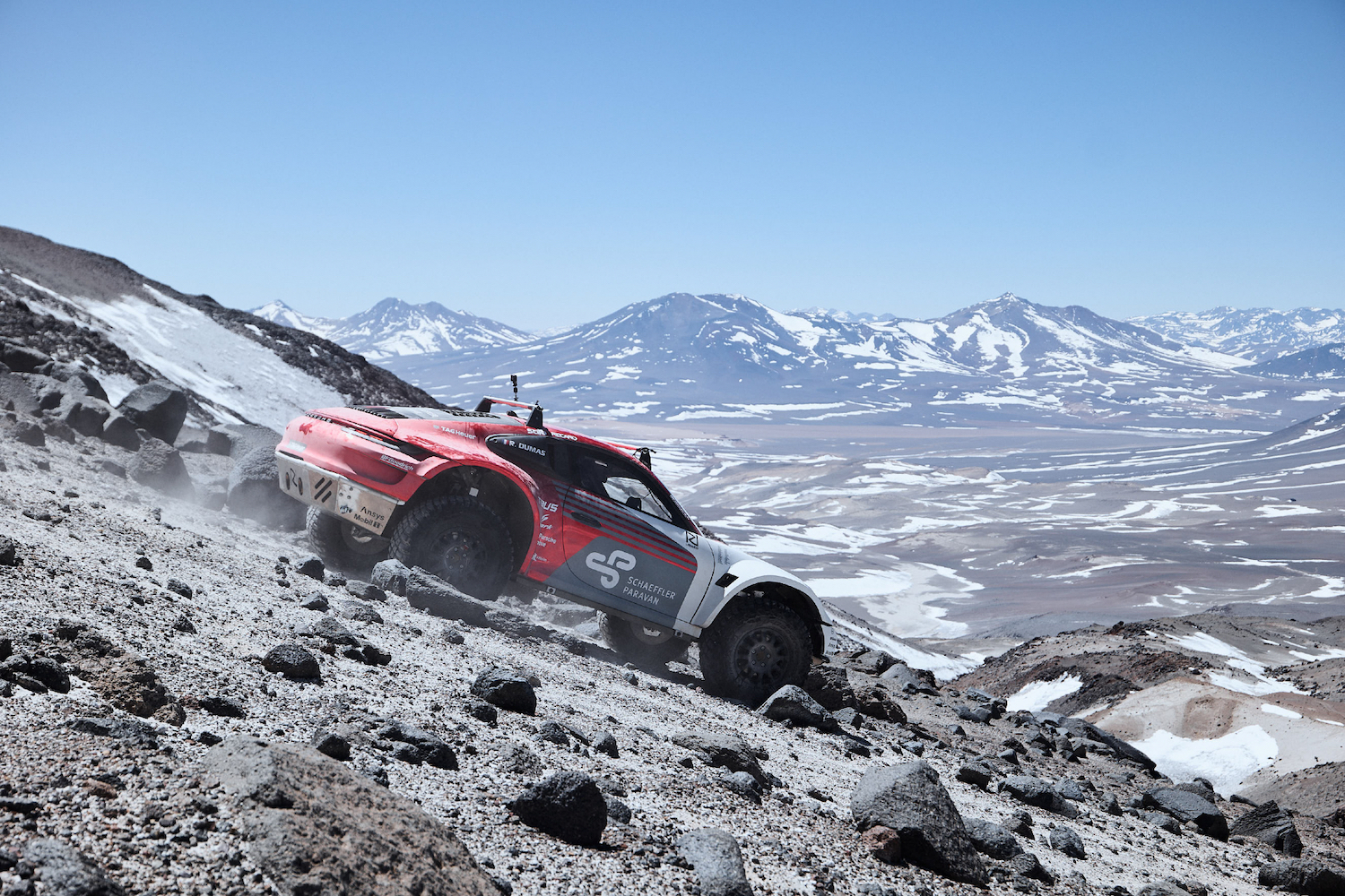 Porsche 911 Dakar Prototype going down a rocky terrain with snow on the ground with mountains in the back.