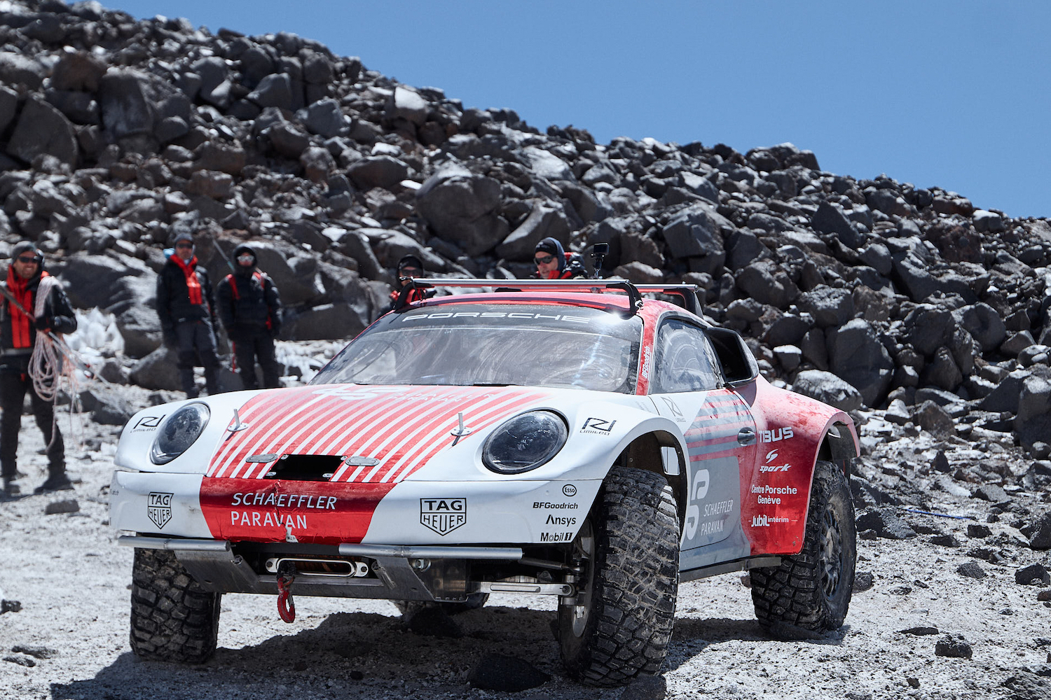 Front end of Porsche 911 Dakar Prototype going up a rocky volcano with blue skies in the back.